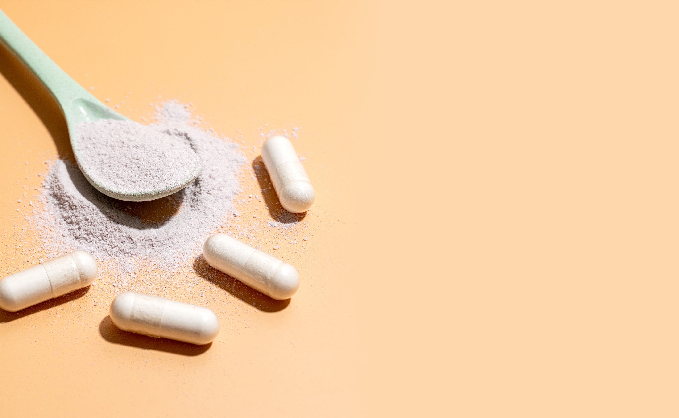 Spoonful of powder surrounded by capsules on a light orange background.