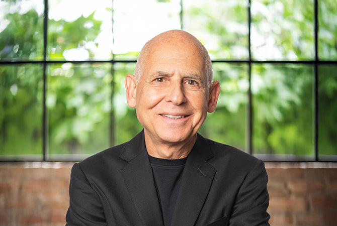 Dr. Daniel Amen in front of a blurred background of a window and green trees in the distance.