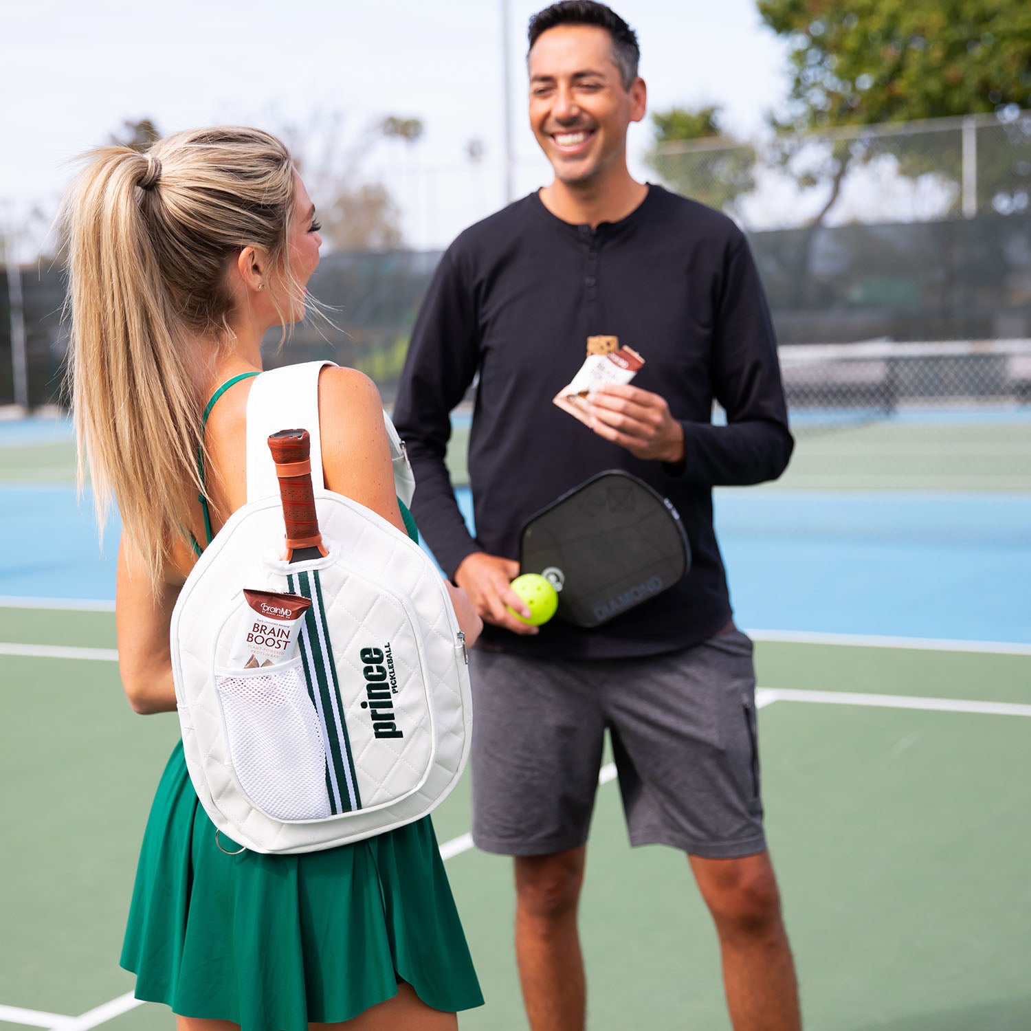 Man and woman conversing on a pickleball court; man is holding a protein bar.