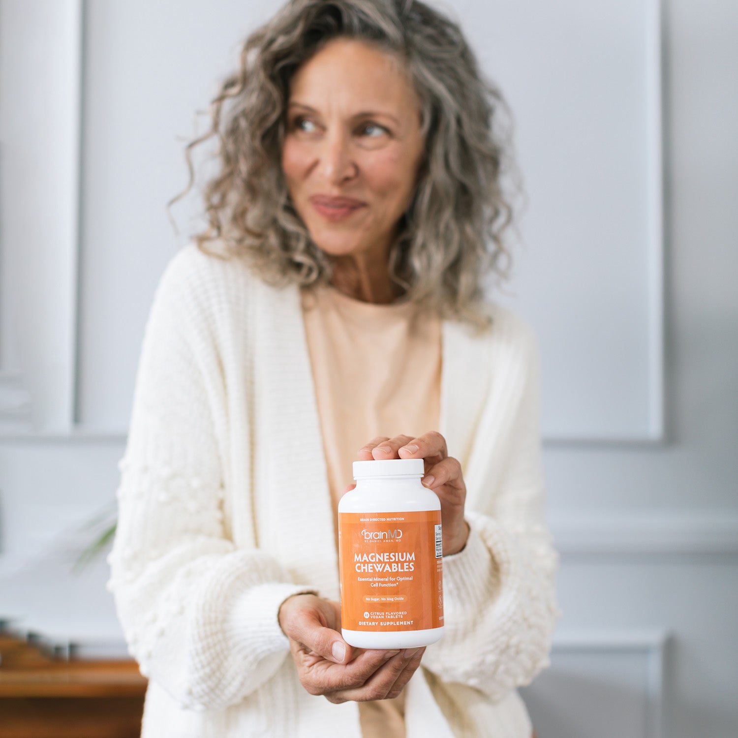 Smiling woman holding an orange supplement bottle against a white wall.