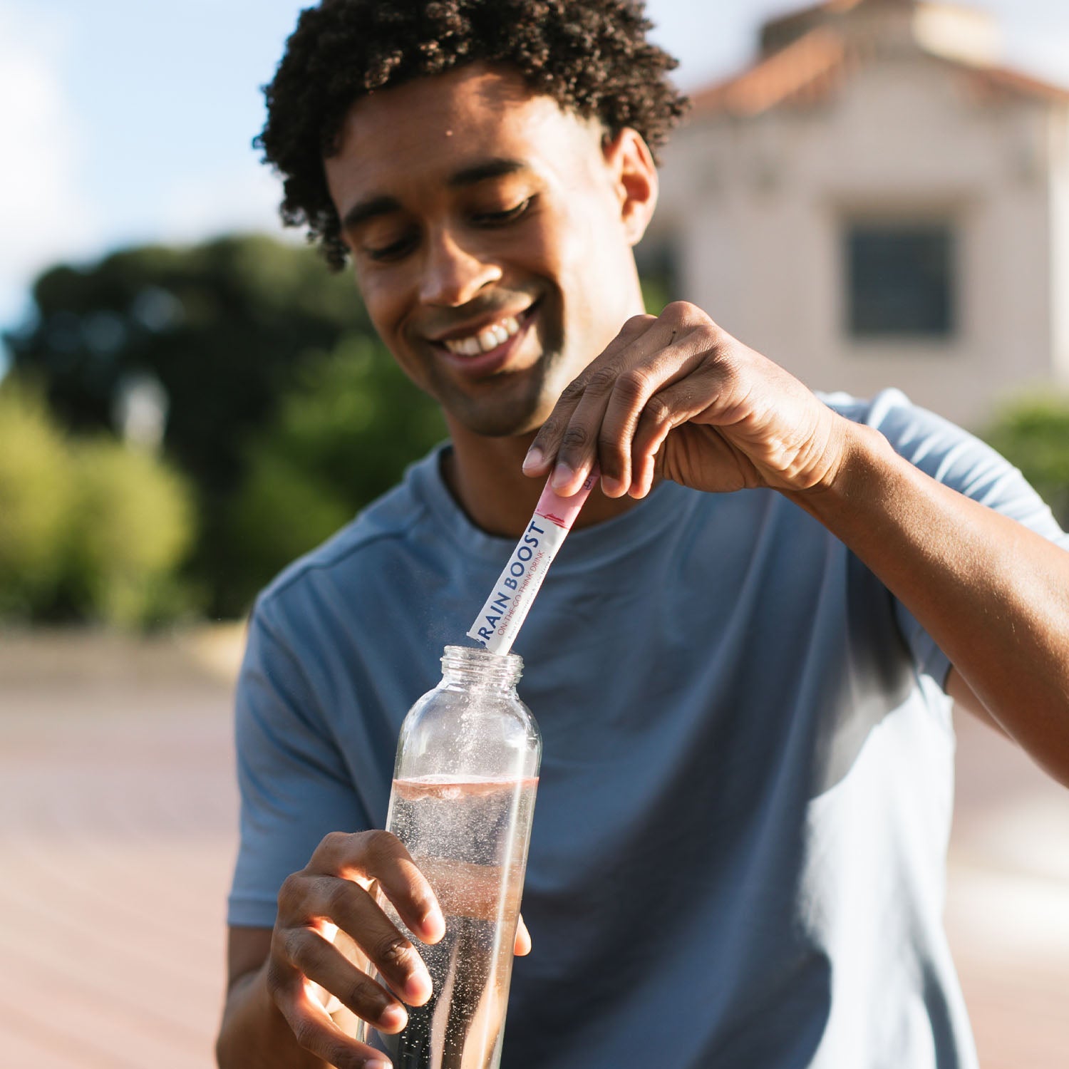 BrainMD Brain Boost On-The-Go: man pouring powder from a packet into a water bottle, blurred exterior background