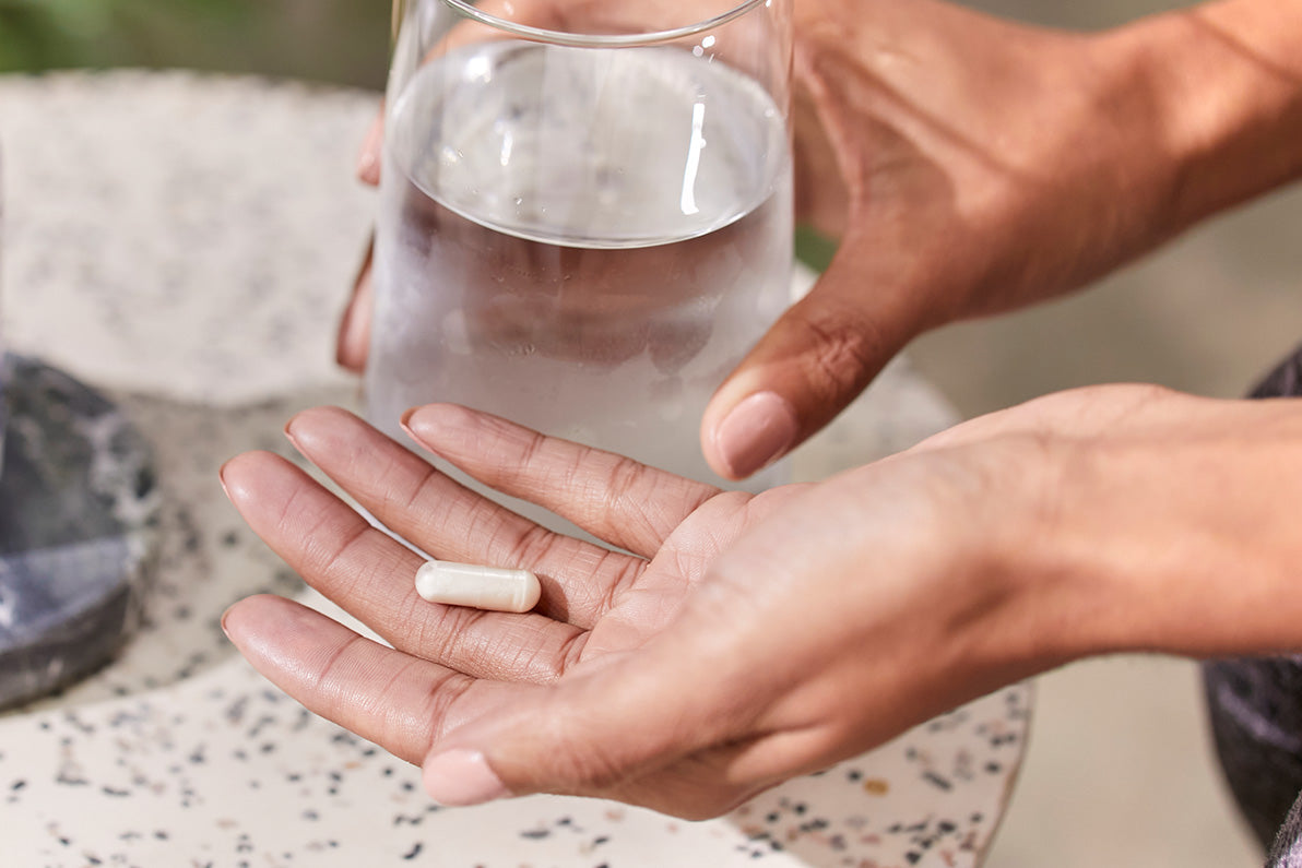 Woman holding a pill in one hand and a glass of water in the other.