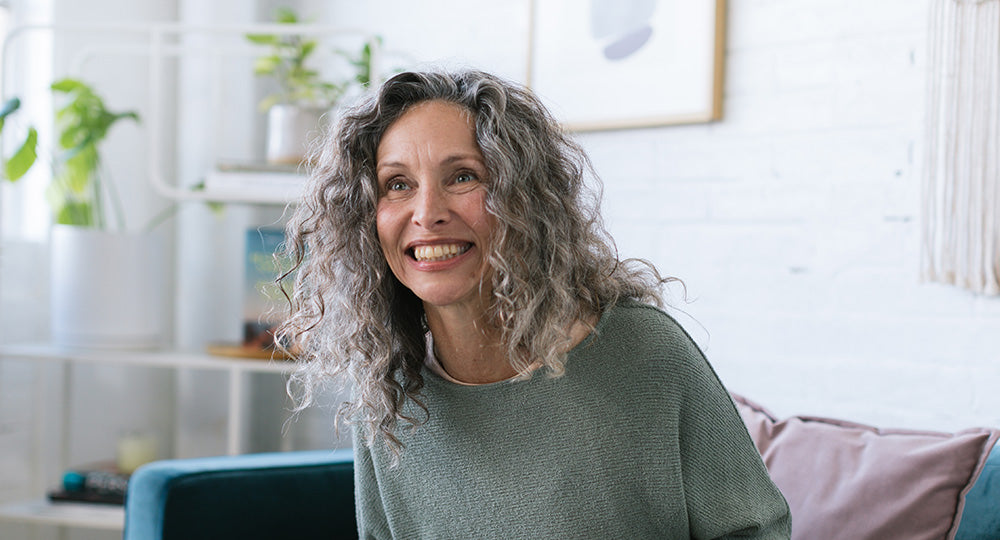 Woman with curly gray hair smiling in a bright living room.