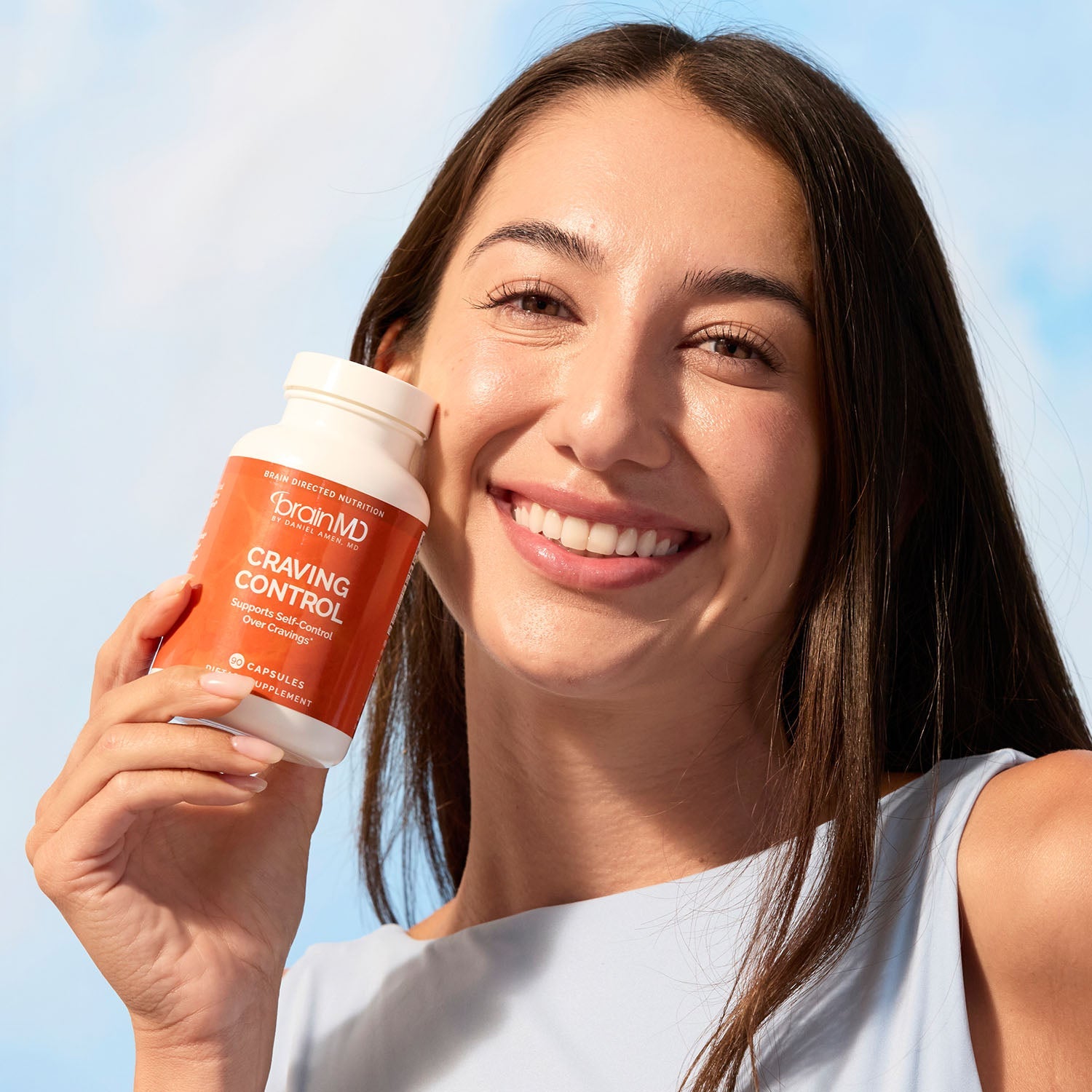 BrainMD Craving Control: smiling woman holding orange supplement bottle against her cheek, blurred sky background