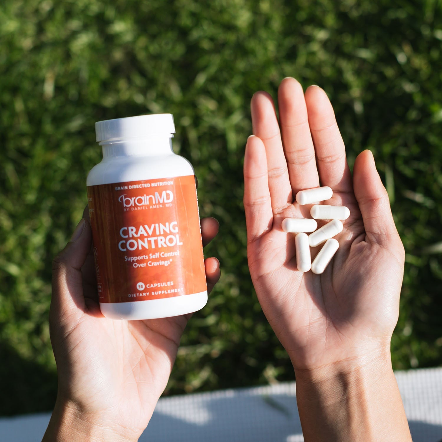 BrainMD Craving Control: person holding orange supplement bottle in one hand & capsules in the other, blurred lawn background
