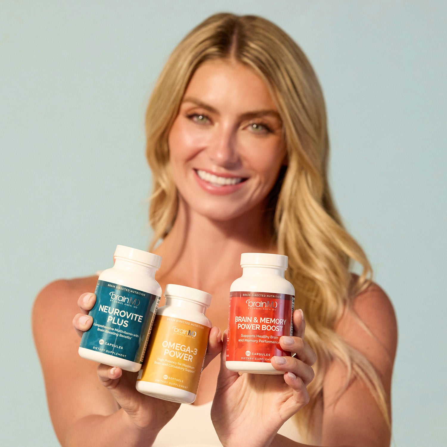 BrainMD Daily Essentials Bundle: woman holds three supplement bottles of various colors against an off-white background