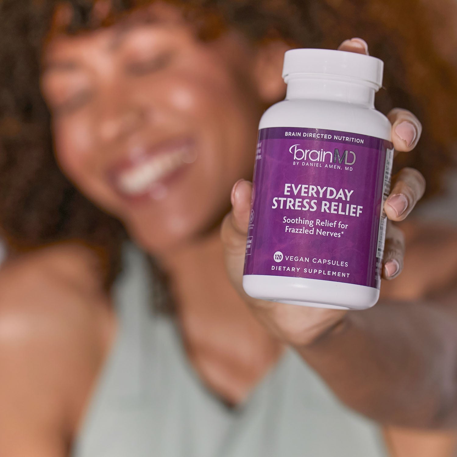 BrainMD Everyday Stress Relief: blurred image of smiling woman in background, holds a purple supplement bottle towards camera