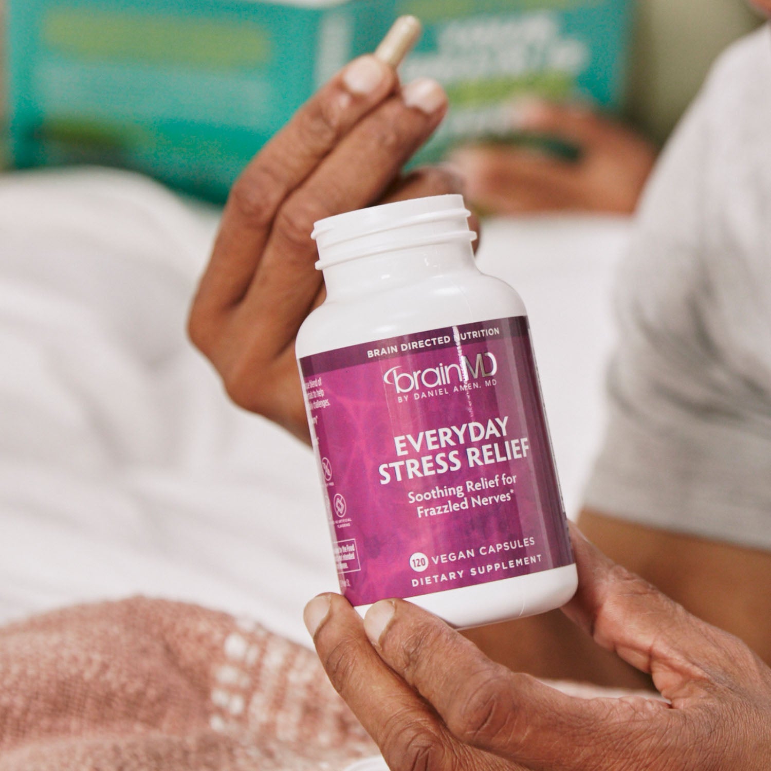 BrainMD Everyday Stress Relief: person holding a purple supplement bottle, blurred bedroom in the background