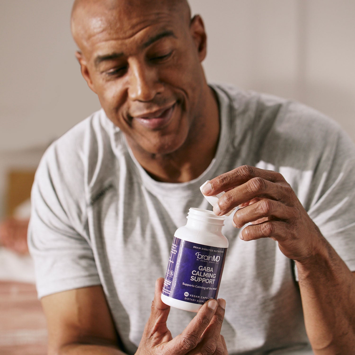 BrainMD GABA Calming Support: smiling man opens purple supplement bottle, blurred interior background