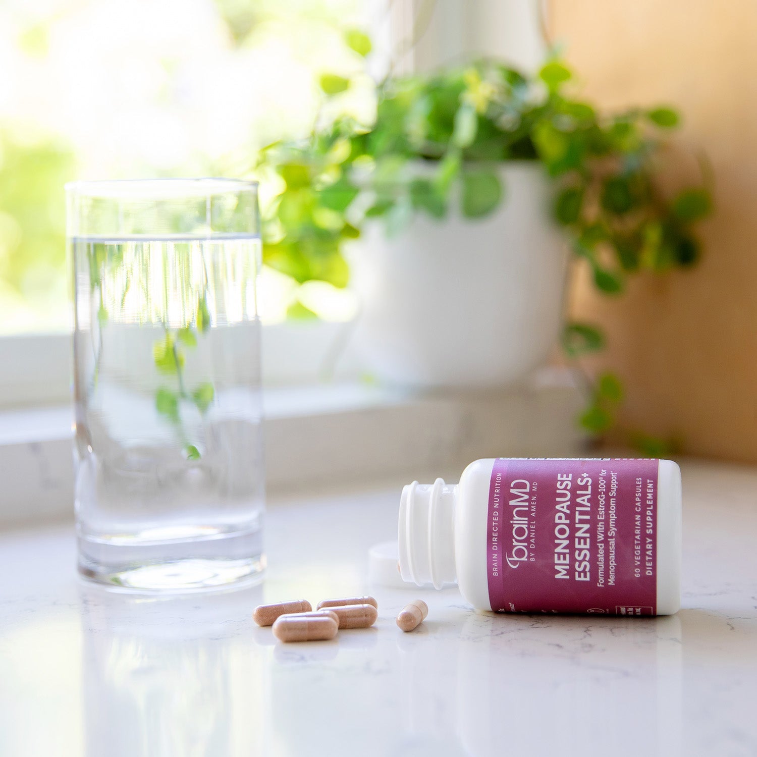 BrainMD Menopause Essentials+: prone supplement bottle flanked by capsules and a glass of water on a kitchen countertop