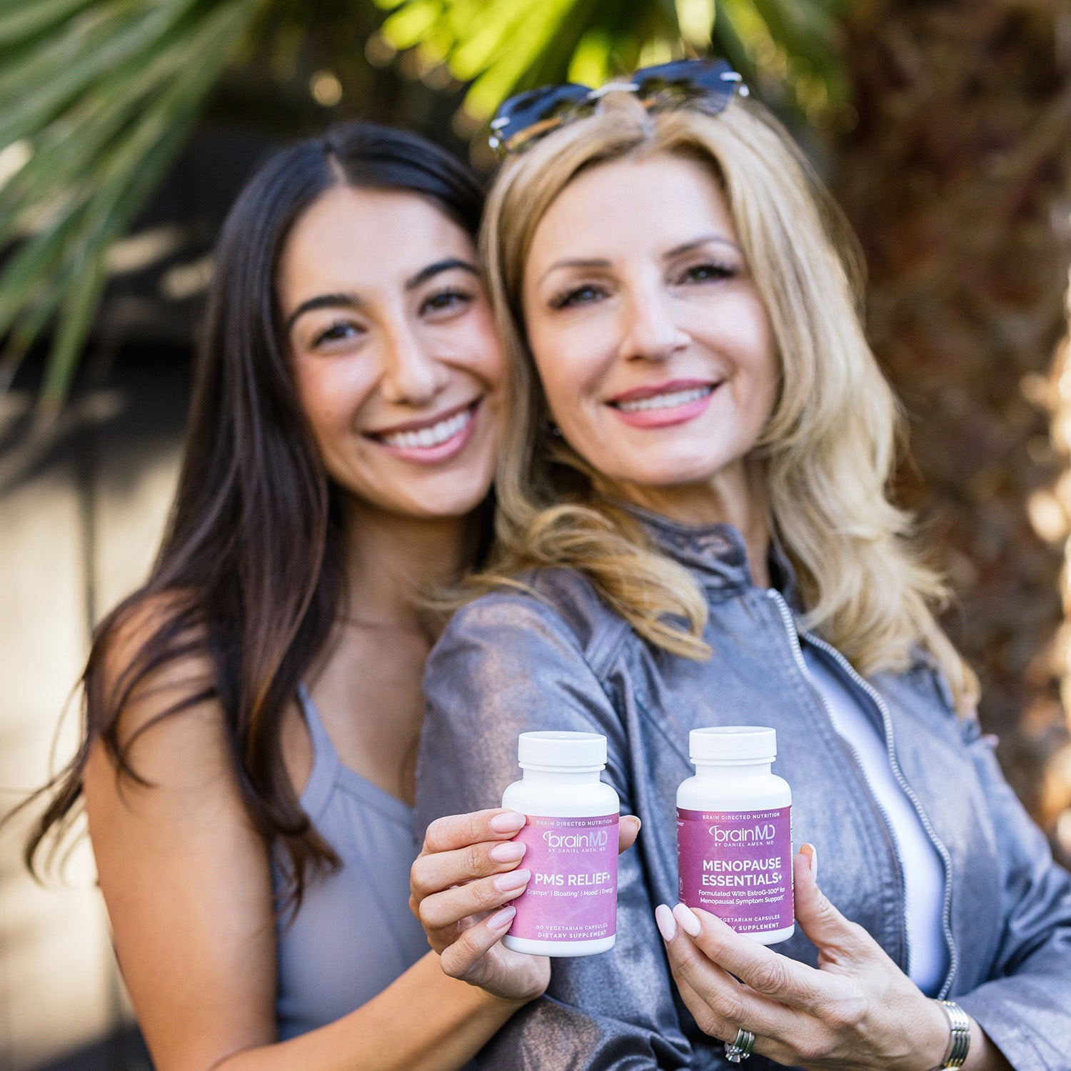 BrainMD Menopause Essentials+: two women hold supplement bottles with a blurred palm tree in the background