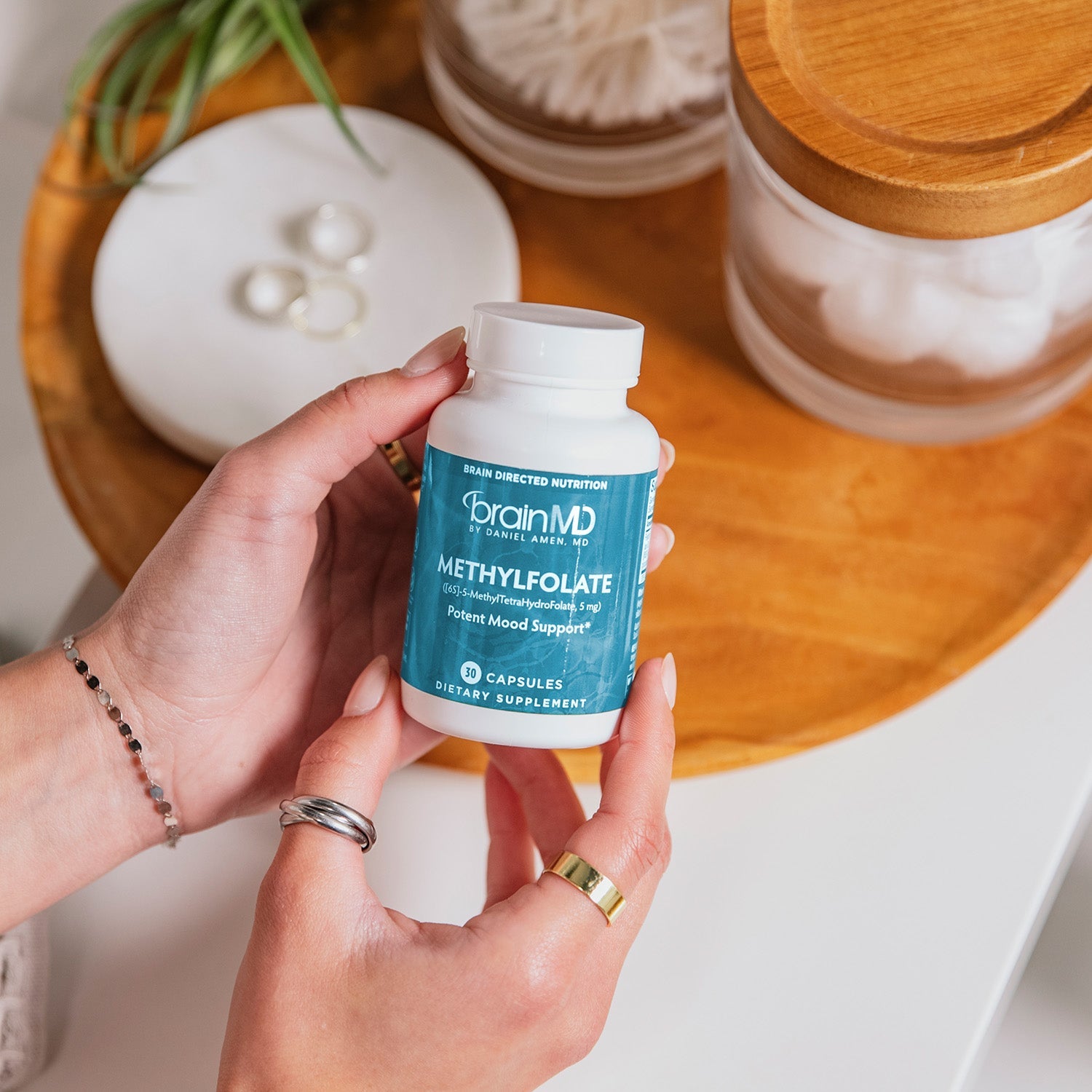 BrainMD MethylFolate: woman holding a blue supplement bottle above a wooden plate and assorted beauty items