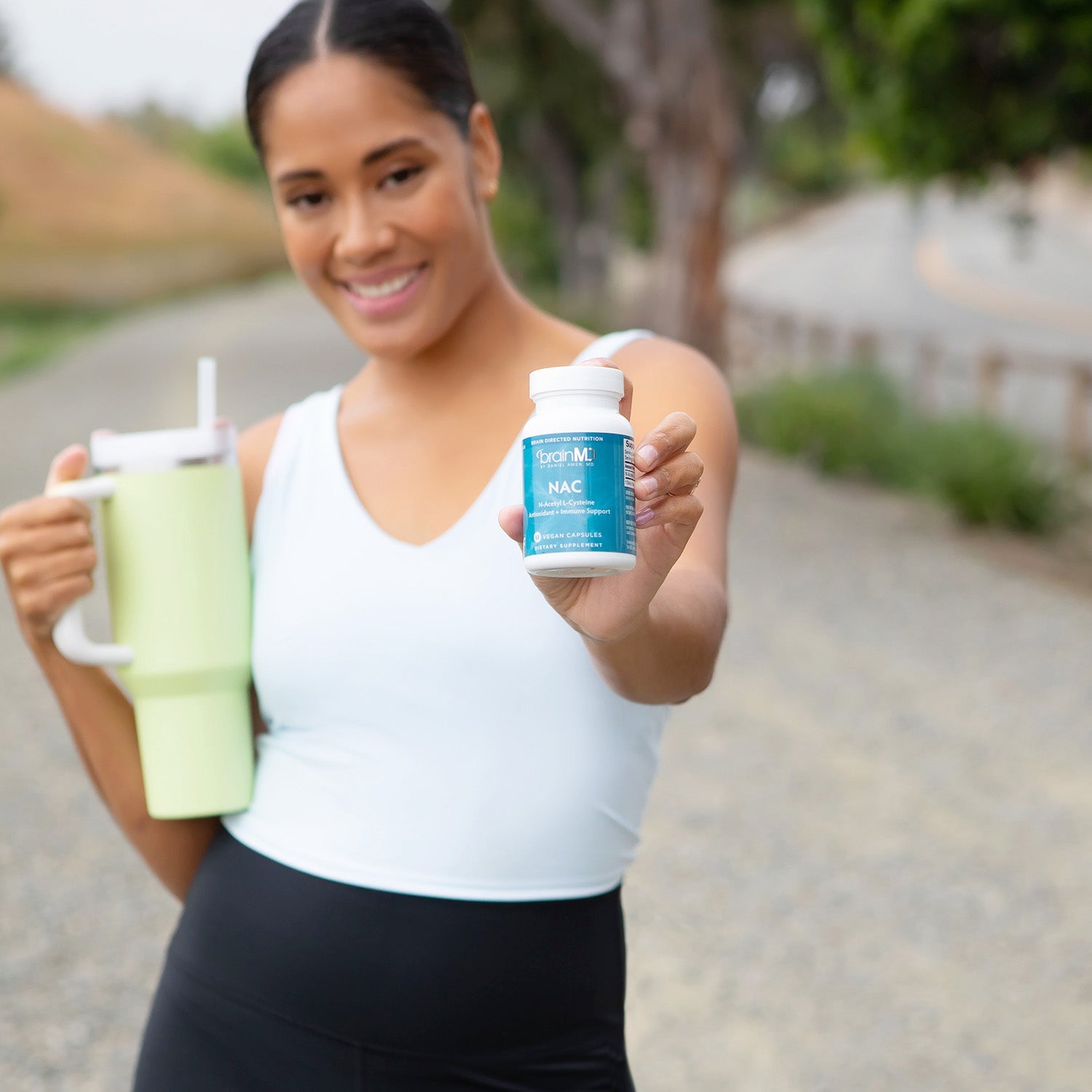 BrainMD NAC (N-Acetyl L-Cysteine): woman on a walking trail holds a NAC bottle in one hand and a water bottle in the other