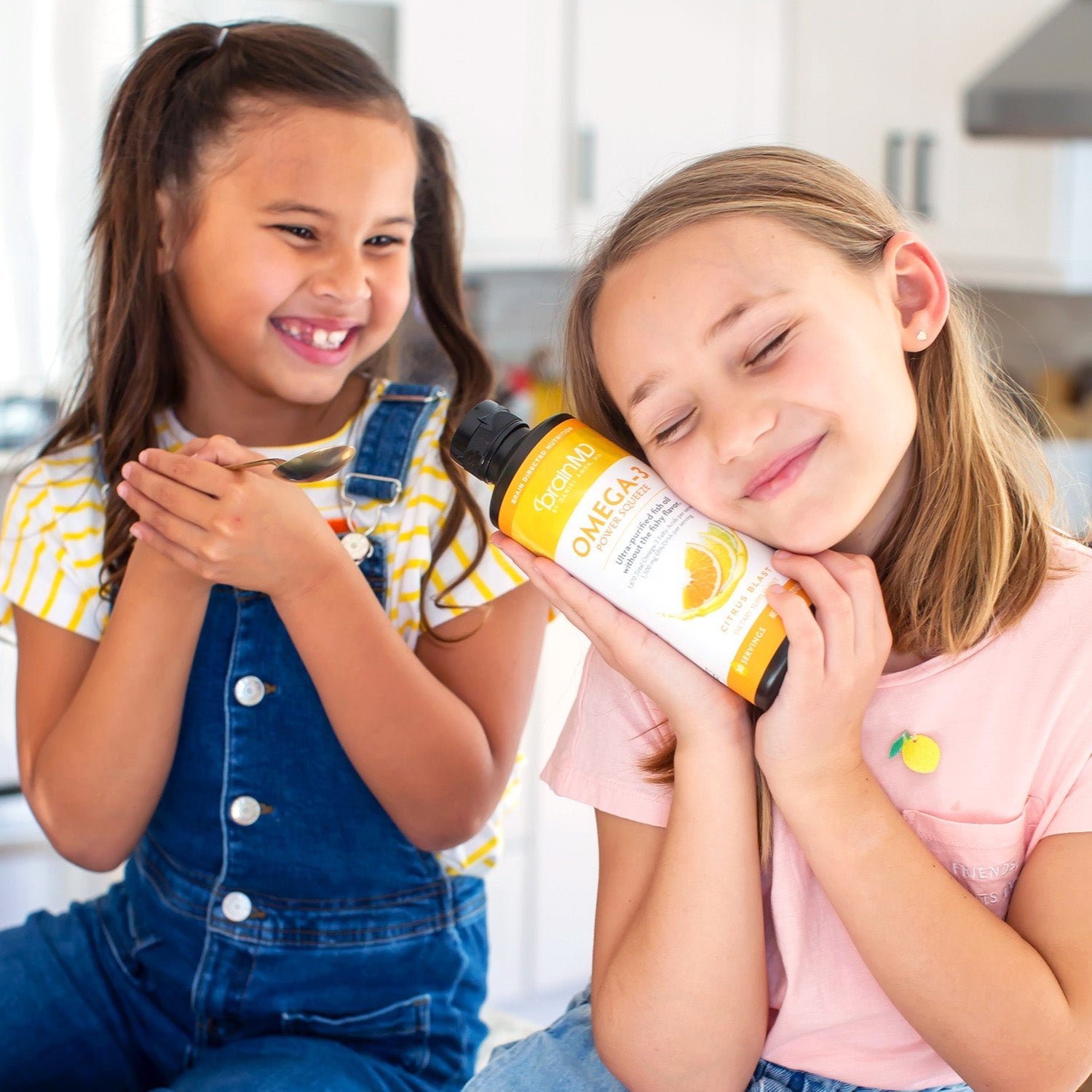 BrainMD Omega-3 Power Squeeze: two girls in a kitchen, one resting her head on an orange and white supplement bottle
