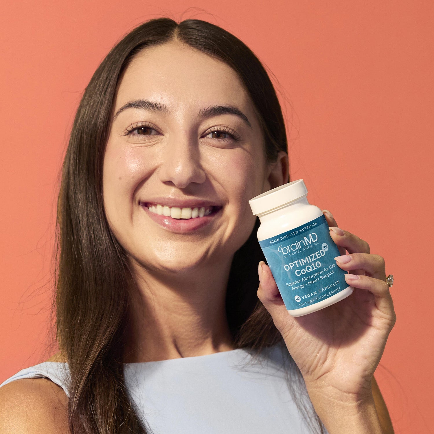 BrainMD Optimized CoQ10: smiling woman holding a blue supplement bottle against an orange background
