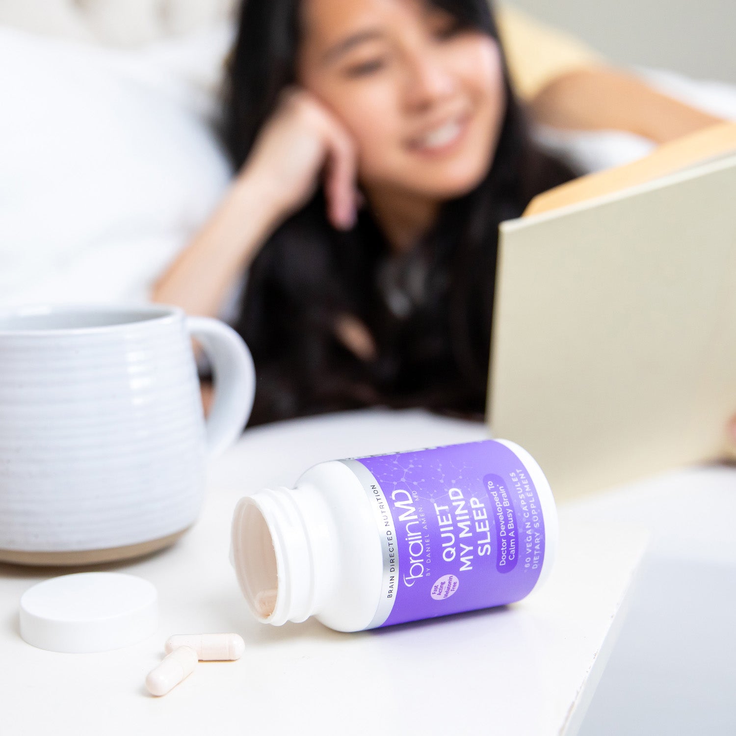 BrainMD Quiet My Mind Sleep: open supplement bottle with capsules on nightstand, smiling woman is blurred in the background