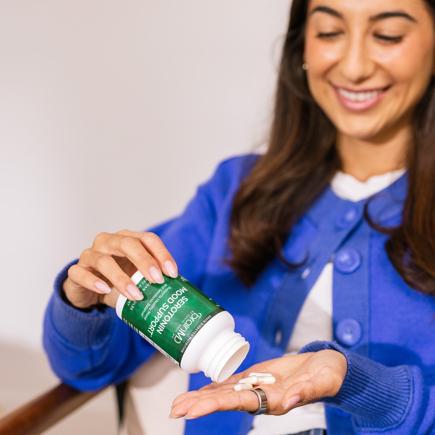 BrainMD Serotonin Mood Support: smiling woman in the background pours capsules into her hand from a green supplement bottle