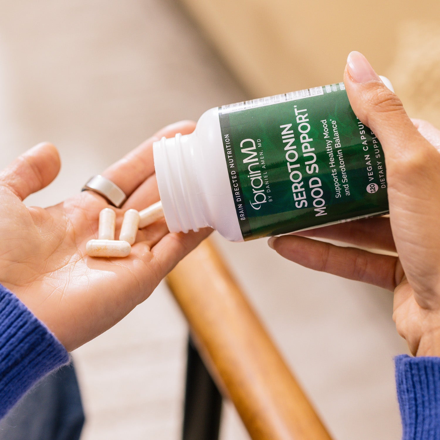 BrainMD Serotonin Mood Support: woman pours capsules into hand from a green supplement bottle, blurred interior background