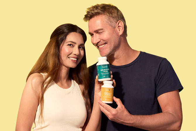 Man and woman holding supplement bottles standing in front of a yellow background.