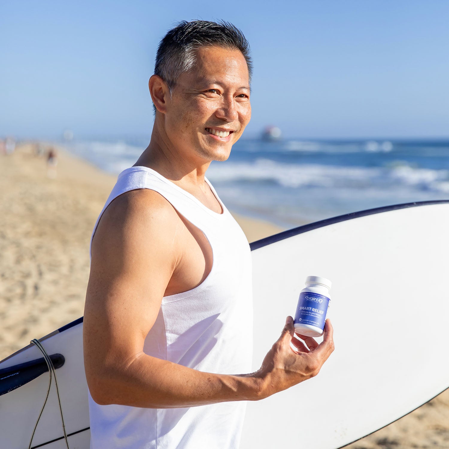 Man on the beach holding a surfboard in one hand and a blue supplement bottle in the other.