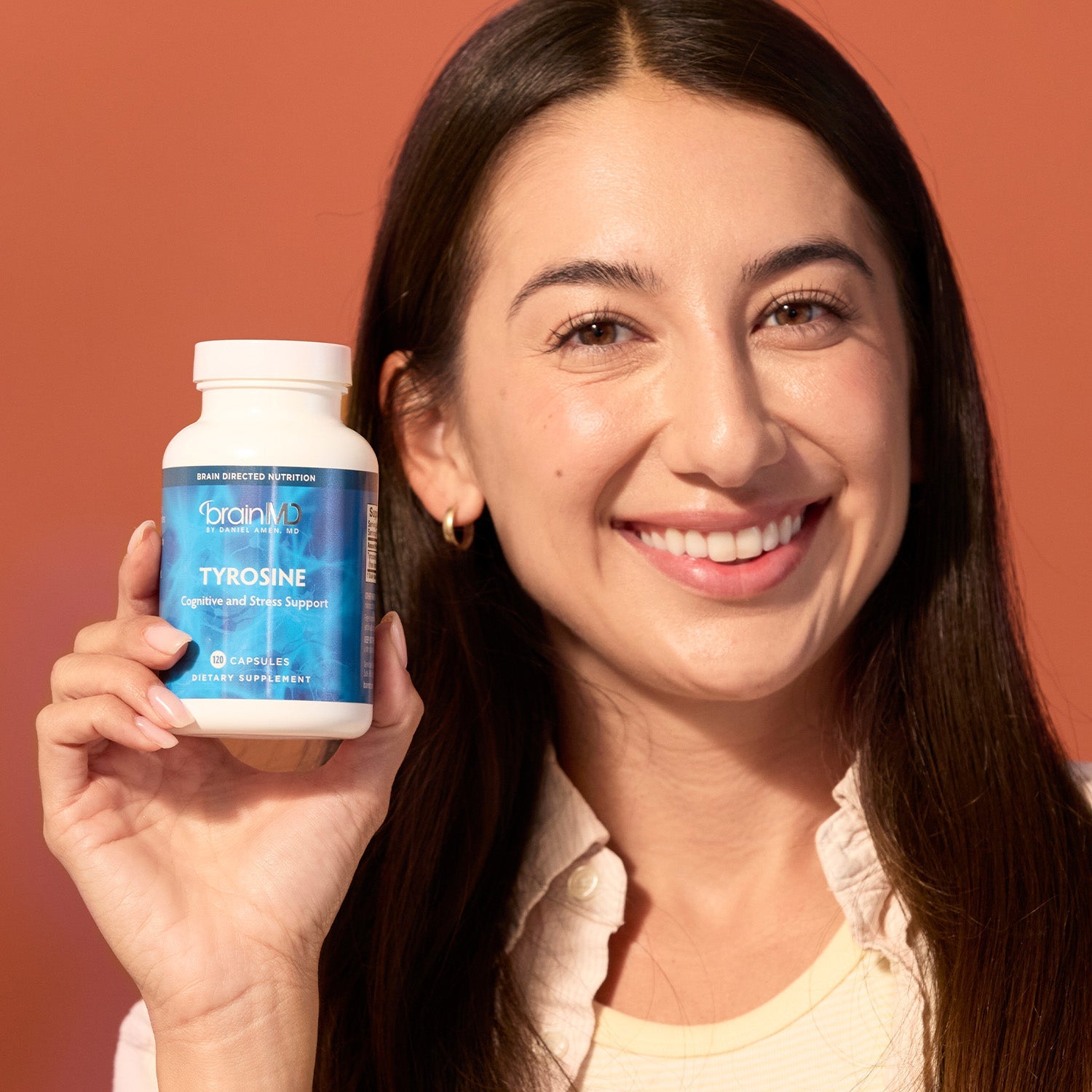 BrainMD Tyrosine: stress support supplement, smiling woman holding supplement bottle next to her face