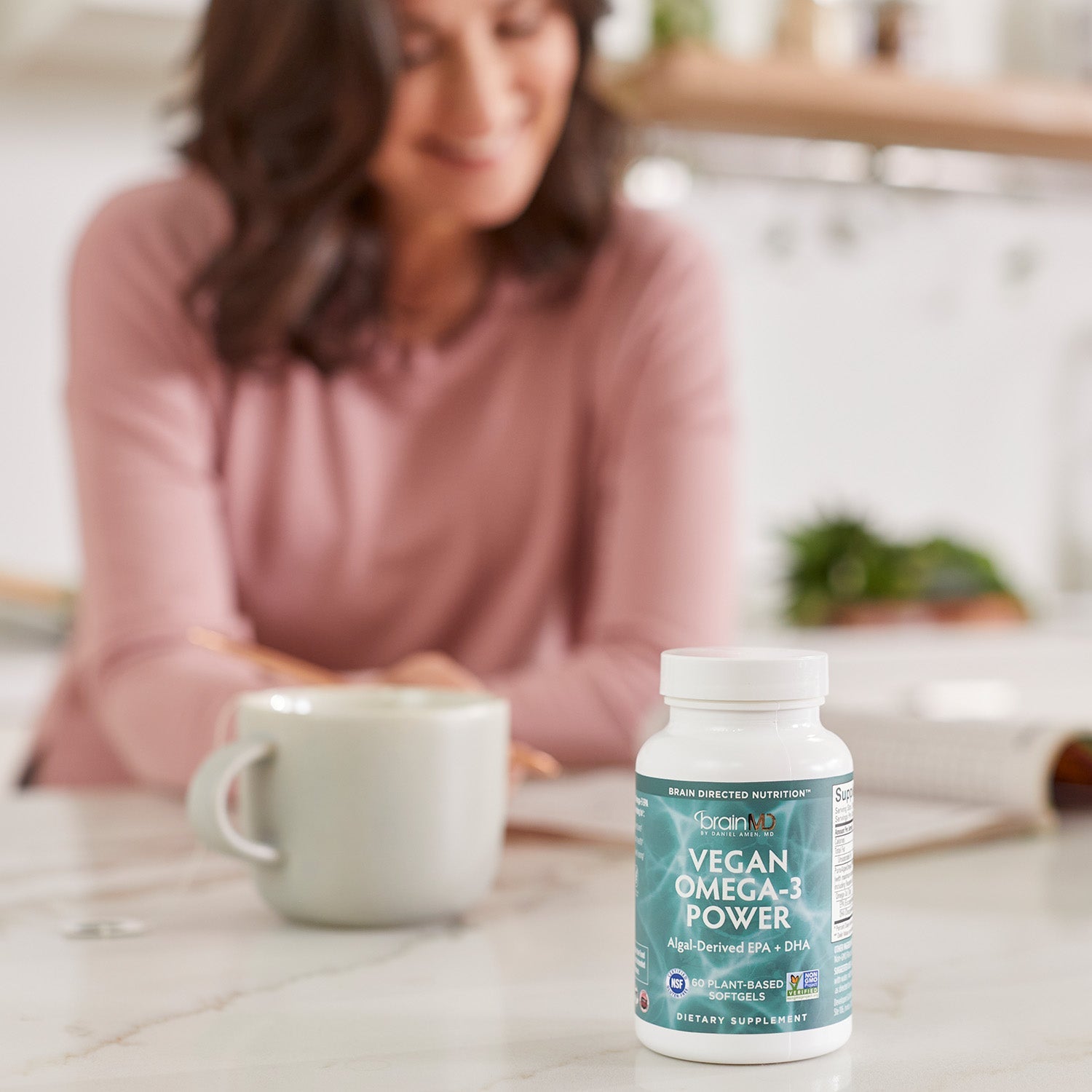 BrainMD Vegan Omega-3 Power: supplement bottle on a kitchen counter in the foreground with a woman blurred in the background