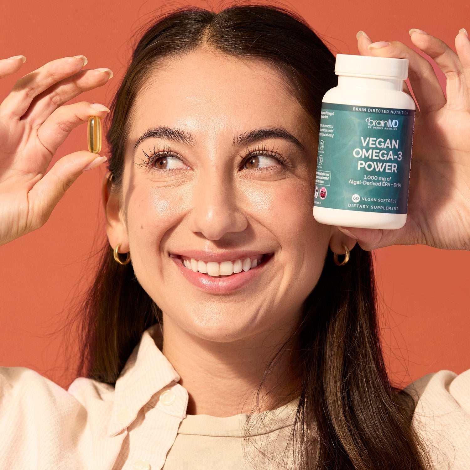 BrainMD Vegan Omega-3 Power: smiling woman holds a softgel in one hand and a green supplement bottle in the other