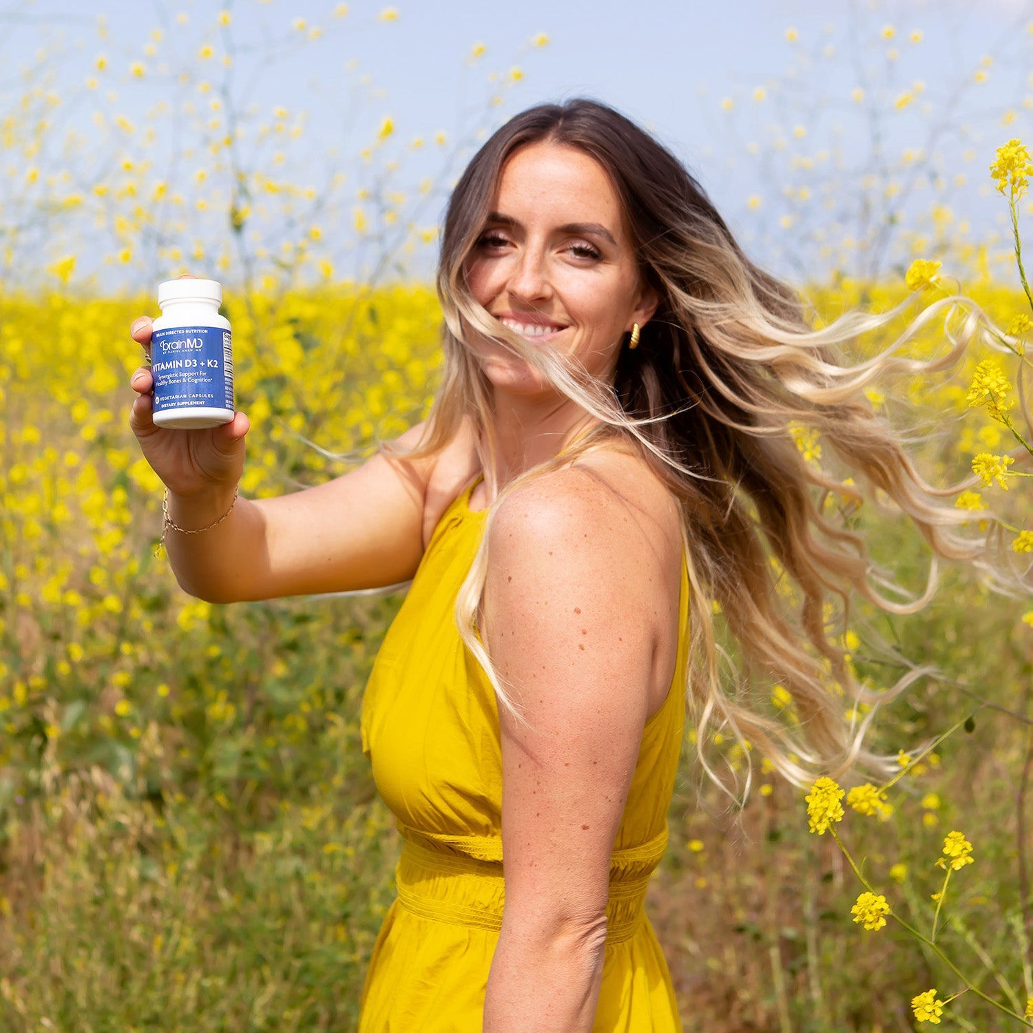 BrainMD Vitamin D3 + K2: woman in yellow dress holds supplement bottle in a field of yellow flowers