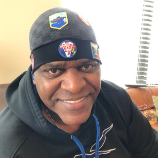 Duval Love wearing a black beanie with patches and a black hoodie, sitting indoors.