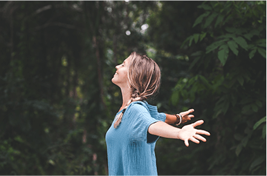 Smiling woman with outstretched arms against a forest background.