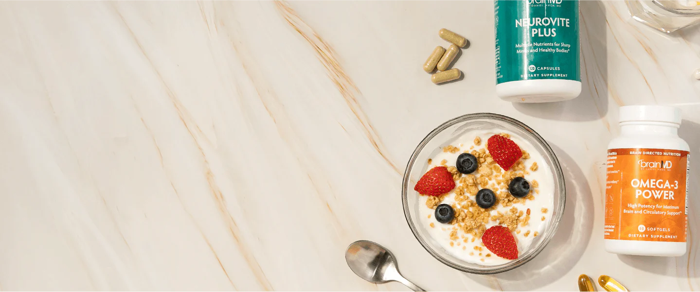 Yogurt bowl with berries and granola next to BrainMD's Neurovite Plus and Omega 3 Power supplement bottles on a light marble surface.